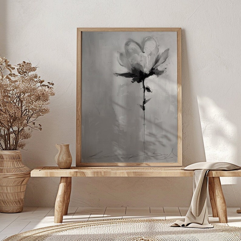 A monochromatic framed canvas print featuring a stylized flower in shades of gray. The flower is depicted with loose brushstrokes, creating a textured and painterly effect. The background is a soft, muted gray, providing a subtle contrast to the darker tones of the flower. The overall style is minimalist and modern, with a focus on the artistic expression of the floral subject. The print is displayed on a light-colored wooden bench, adding to the serene and calming atmosphere of the scene.