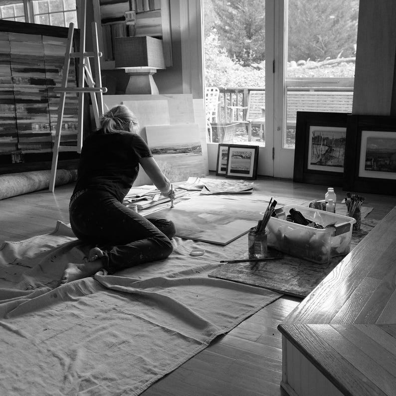Artist Sallie Otenasek at work in studio with finished pieces in the background.  Sallie is sitting on the floor, she prefers to work that way rather than standing at an easel.
