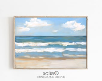 May include: A framed beach scene painting. The artwork depicts a blue ocean with white-capped waves under a light blue sky with fluffy white clouds. The sandy beach is painted in shades of tan and beige.