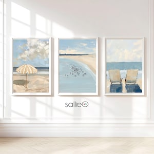 May include: Three framed paintings of beach scenes. The leftmost painting shows a yellow and white striped beach umbrella on a sandy beach. The middle painting shows a blue ocean with a sandy beach and a group of birds in the water. The rightmost painting shows two beach chairs on a sandy beach with a blue ocean in the background.