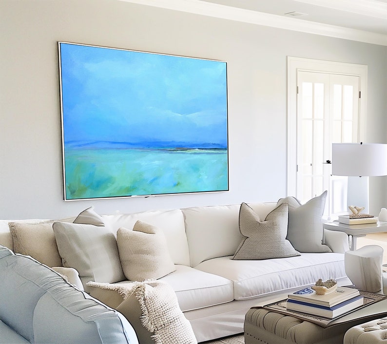 May include: A white sectional sofa with light blue throw pillows and a white throw blanket. The sofa is in front of a large abstract painting of a blue sky and green water. A small table with books and a decorative object sits in front of the sofa.