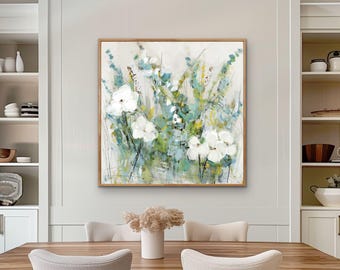 May include: A framed floral painting featuring white flowers with green and blue accents. The artwork is displayed in a dining room setting, with a wooden table and chairs visible in the foreground. The painting has a light, airy feel.