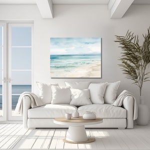 A watercolor painting of a beach scene with blue water, white sand, and a light blue sky with white clouds. The painting is hanging on a white wall in a living room setting with a white couch and a round coffee table.