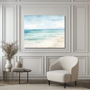 A watercolor painting of a seascape with blue water, white sand, and a light blue sky. The painting is framed and hanging on a white wall.