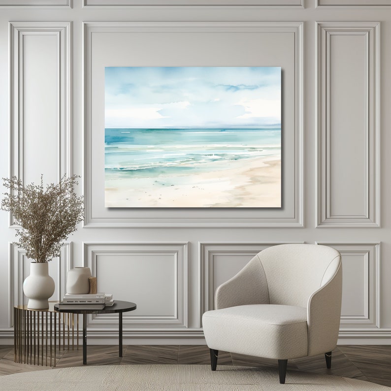 A watercolor painting of a seascape with blue water, white sand, and a light blue sky. The painting is framed and hanging on a white wall.