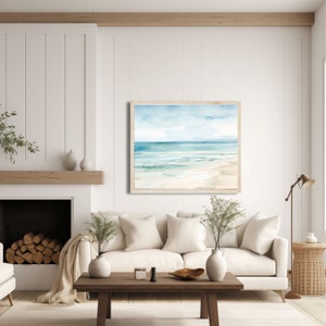 A framed watercolor painting of a beach scene with blue water, white sand, and a light blue sky. The painting is hanging on a white wall in a living room setting. The painting is framed in a light wood frame.
