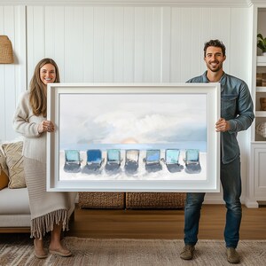 May include: A framed art print depicting six beach chairs on a sandy shore under a cloudy sky. The chairs are various shades of blue and turquoise. The artwork is held by a man and a woman.