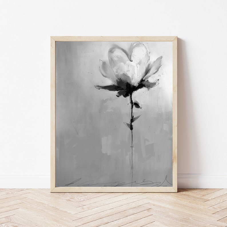 A framed grayscale abstract painting of a single flower. The flower is rendered in shades of gray and black, with visible brushstrokes. The background is a textured gray canvas. The painting has a minimalist aesthetic and evokes a sense of serenity. The frame is light wood. The artwork is suitable for modern or contemporary home decor.