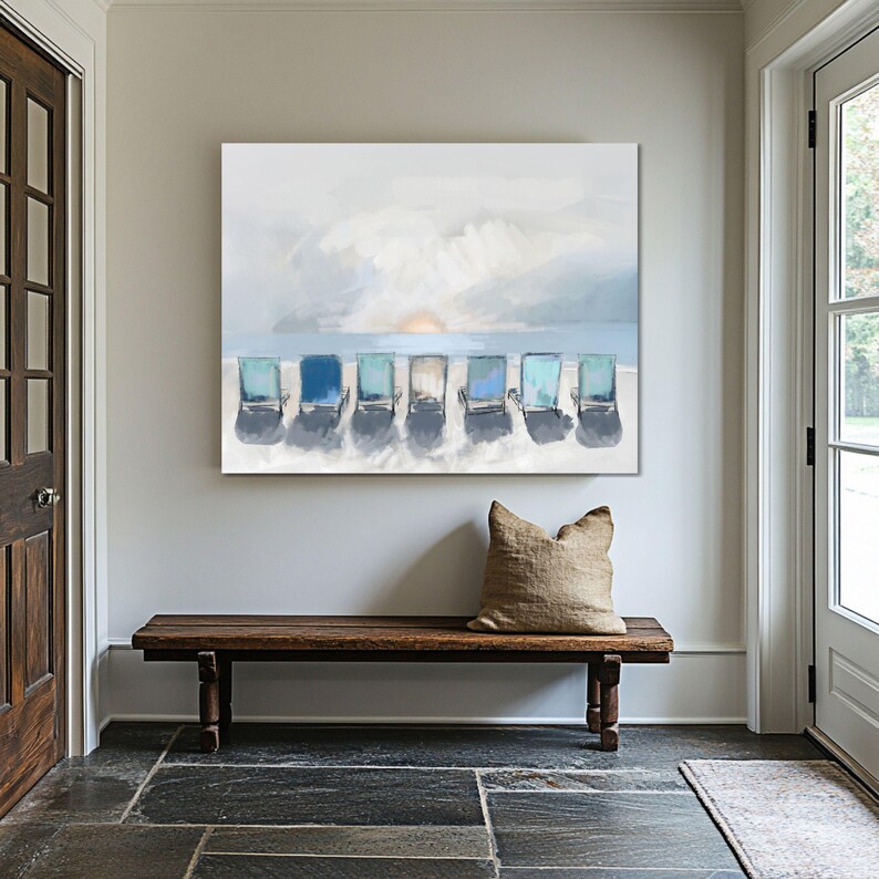 May include: An abstract painting depicts seven beach chairs in varying shades of blue, arranged on a sandy shore. The background features a soft, impressionistic sky with a setting sun. The artwork is displayed above a wooden bench.