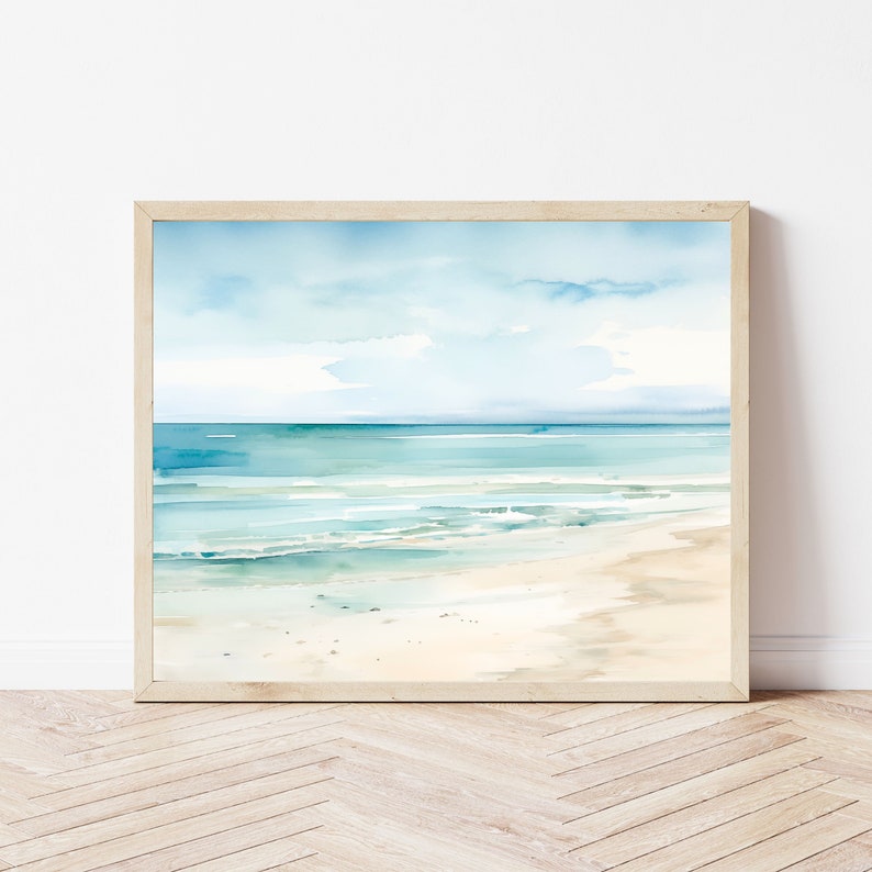 A framed watercolor painting of a beach scene. The painting features a light blue sky with white clouds, a turquoise ocean with white waves, and a sandy beach. The frame is light wood.