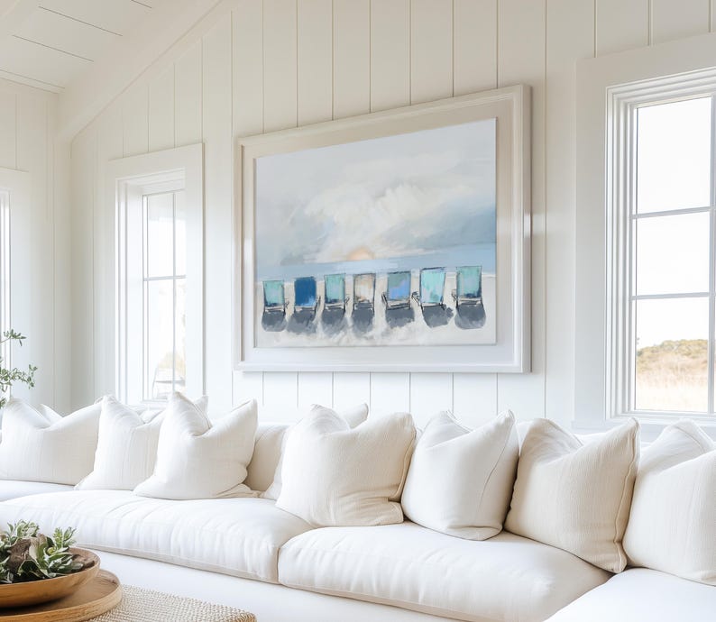 May include: A large framed painting depicts a beach scene with several blue and turquoise beach chairs on a sandy shore under a cloudy sky. The painting hangs above a white sofa with several white pillows, creating a coastal theme.