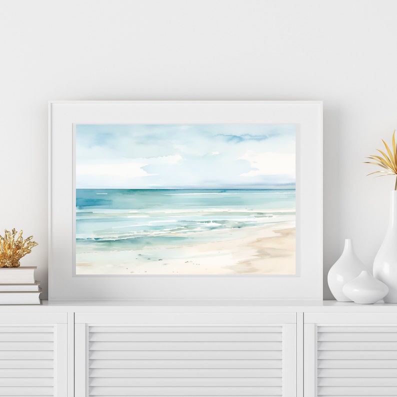 A framed watercolor painting of a beach scene with blue water, white sand, and a light blue sky with clouds.