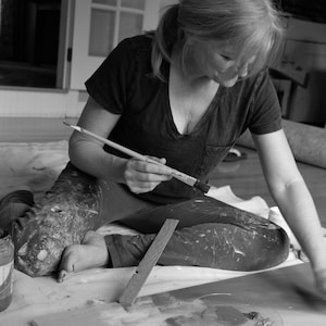 May include: A woman sits on the floor, wearing paint-splattered clothing, and holds a paintbrush in her hand. She is working on a painting project, with a paint can and a wooden board nearby.