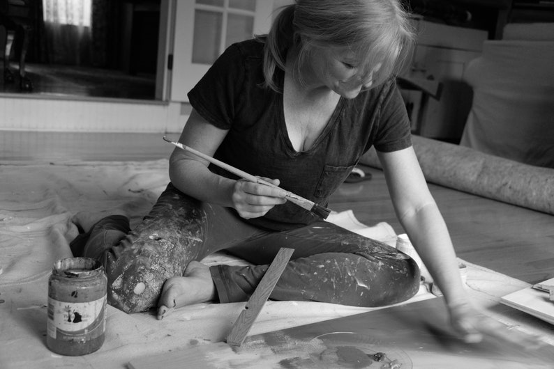 May include: A woman sits on the floor, wearing paint-splattered clothing, and holds a paintbrush in her hand. She is working on a painting project, with a paint can and a wooden board nearby.