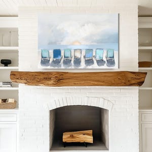 May include: A canvas print depicting a beach scene with six blue and turquoise beach chairs lined up on the sand, facing the ocean. The sky is a blend of light blue and white, with a hint of the sun setting on the horizon. The artwork is displayed above a wooden mantel.