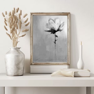 A framed grayscale painting of a single magnolia flower on a textured background. The flower is rendered in shades of gray and black, with subtle brushstrokes suggesting texture and movement. The frame is light-colored, and the overall style is minimalist and elegant. The painting is displayed on a white shelf with a vase of dried flowers and a book.