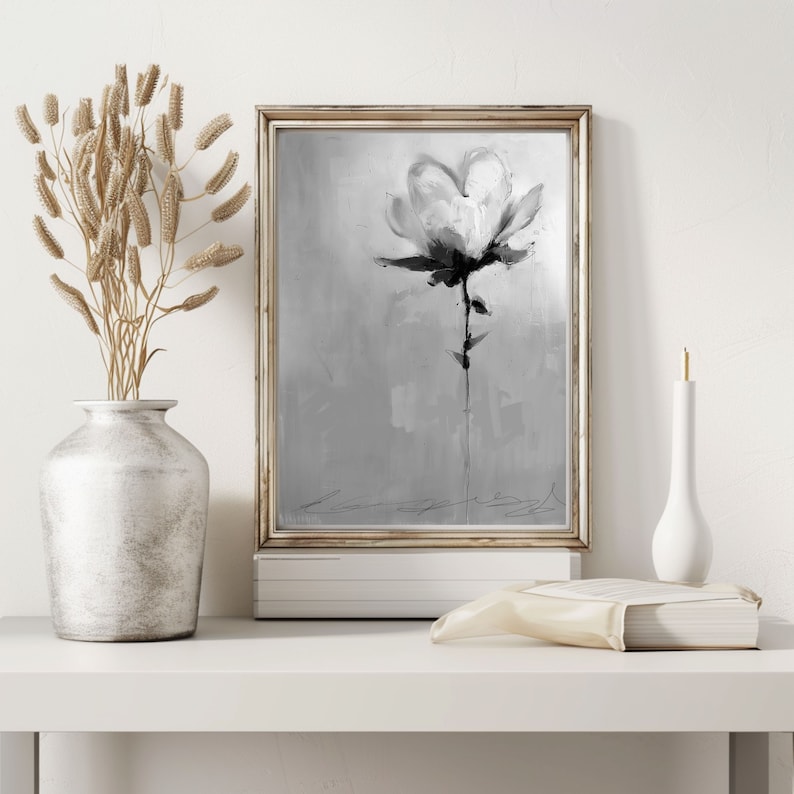 A framed grayscale painting of a single magnolia flower on a textured background. The flower is rendered in shades of gray and black, with subtle brushstrokes suggesting texture and movement. The frame is light-colored, and the overall style is minimalist and elegant. The painting is displayed on a white shelf with a vase of dried flowers and a book.