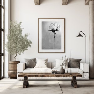A monochrome painting of a single flower, rendered in shades of gray and black, hangs on a white wall above a white couch. The couch features gray and white pillows. A rustic wooden coffee table sits in front of the couch, and a small olive tree in a wooden pot is placed to the left. A floor lamp with a black shade stands in the corner. The overall aesthetic is minimalist and modern.