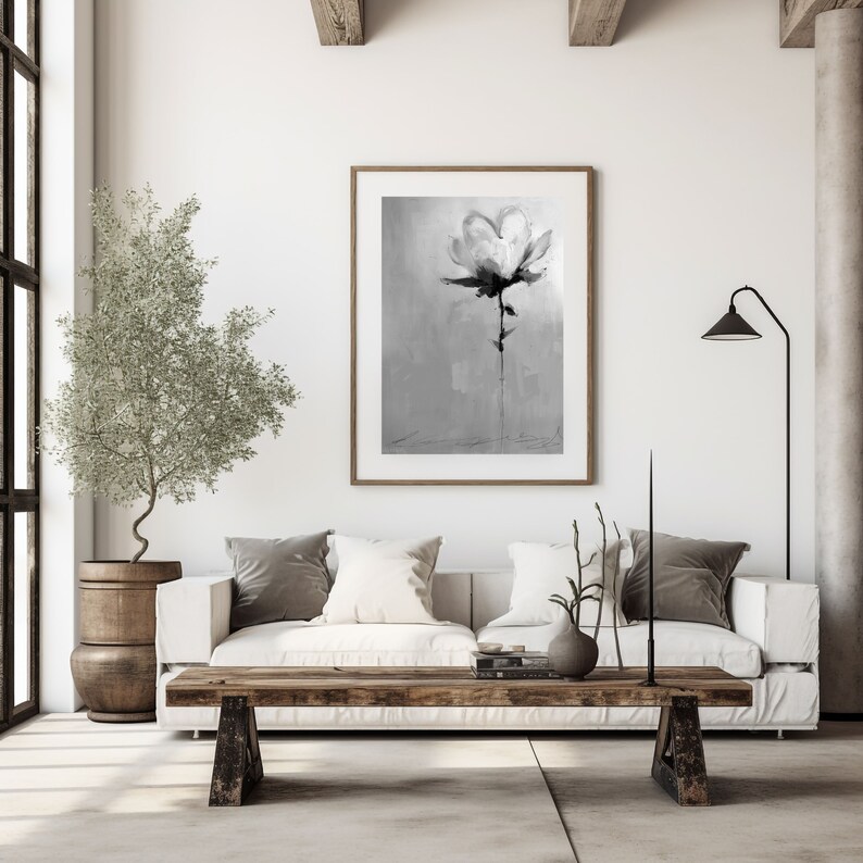 A monochrome painting of a single flower, rendered in shades of gray and black, hangs on a white wall above a white couch. The couch features gray and white pillows. A rustic wooden coffee table sits in front of the couch, and a small olive tree in a wooden pot is placed to the left. A floor lamp with a black shade stands in the corner. The overall aesthetic is minimalist and modern.