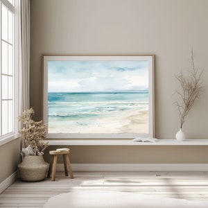 A framed watercolor painting of a beach scene with blue water, white sand, and a cloudy sky. The painting is hanging on a white wall above a white shelf. There is a small wooden stool and a vase with dried branches in front of the painting.