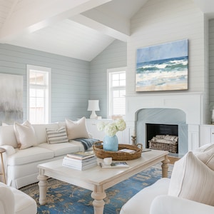 May include: A white living room with a white couch, a coffee table, and a fireplace. The fireplace has a painting of a beach scene above it. The room is decorated in a coastal style with blue and white accents.