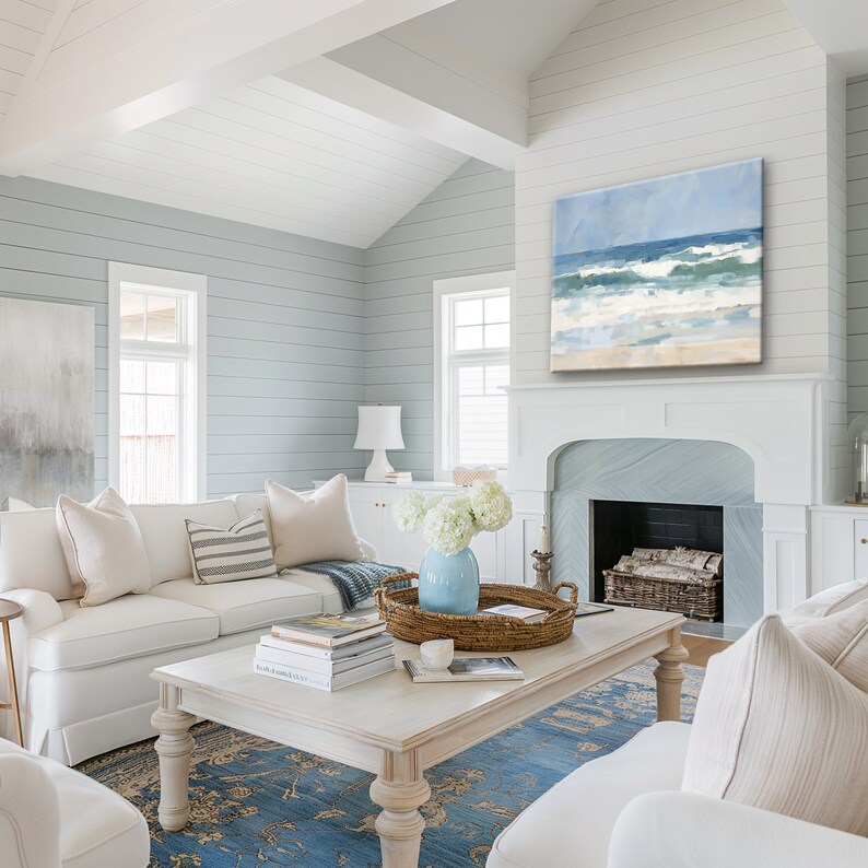 May include: A white living room with a white couch, a coffee table, and a fireplace. The fireplace has a painting of a beach scene above it. The room is decorated in a coastal style with blue and white accents.
