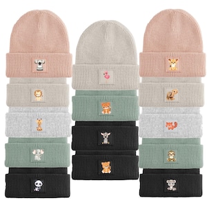 May include: A collection of ribbed knit beanies in shades of pink, grey, and black. Each beanie has a small square patch with an embroidered animal design, like a koala, flamingo, or tiger. The hats are stacked, displaying the range of colours and animal motifs available.