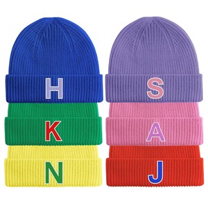 May include: Six ribbed knit beanies in a stack, each in a different colour: blue, green, yellow, purple, pink, and red. Each beanie has a single letter: H, K, N, S, A, and J, in a contrasting colour.