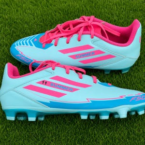 May include: A pair of light blue football boots with bright pink laces, interior lining, and three-stripe branding. The side of the shoe features the text "SUMMER" and the number "11" in pink, with "F50" printed in pink near the toe and heel.
