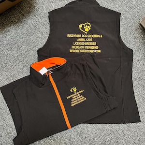 May include: Black dog grooming and animal care uniform set. The set includes a zip-up jacket with an orange zipper and collar, and a sleeveless vest. Both pieces have gold text and a paw print logo. The text reads "RUDDYPAWS DOG GROOMING & ANIMAL CARE".
