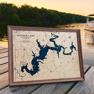 May include: A framed map of Monroe Lake, Bloomington, Indiana. The map shows the lake's outline in dark blue against a light wood backdrop. The frame is a dark brown colour. The text "Monroe Lake Bloomington Indiana" is visible on the map.