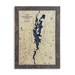 May include: A framed wooden map of Lake Champlain, with the lake in dark blue and surrounding areas in light brown. The map includes text indicating the lake's name, location in the United States and Canada, and other details. The frame is a weathered grey.