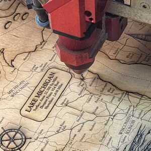 A red laser cutter is cutting a map of Michigan, with the text "LAKEMIGAN" visible on the map.