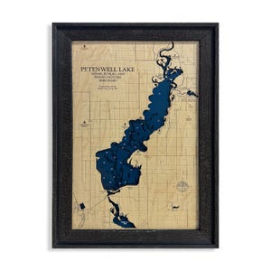 May include: Framed wooden map of Petenwell Lake in Adams, Juneau, and Wood Counties, Wisconsin. The lake is depicted in blue against a light brown background, with depth contours and surrounding land details. The map is encased in a black frame.