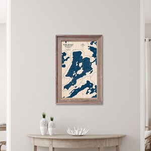 May include: A framed wooden map of Poplar Bay, with a detailed blue depiction of the lake's contours. The map is set against a light-colored background, with the text "Poplar Bay" at the top. The frame is a light brown color.
