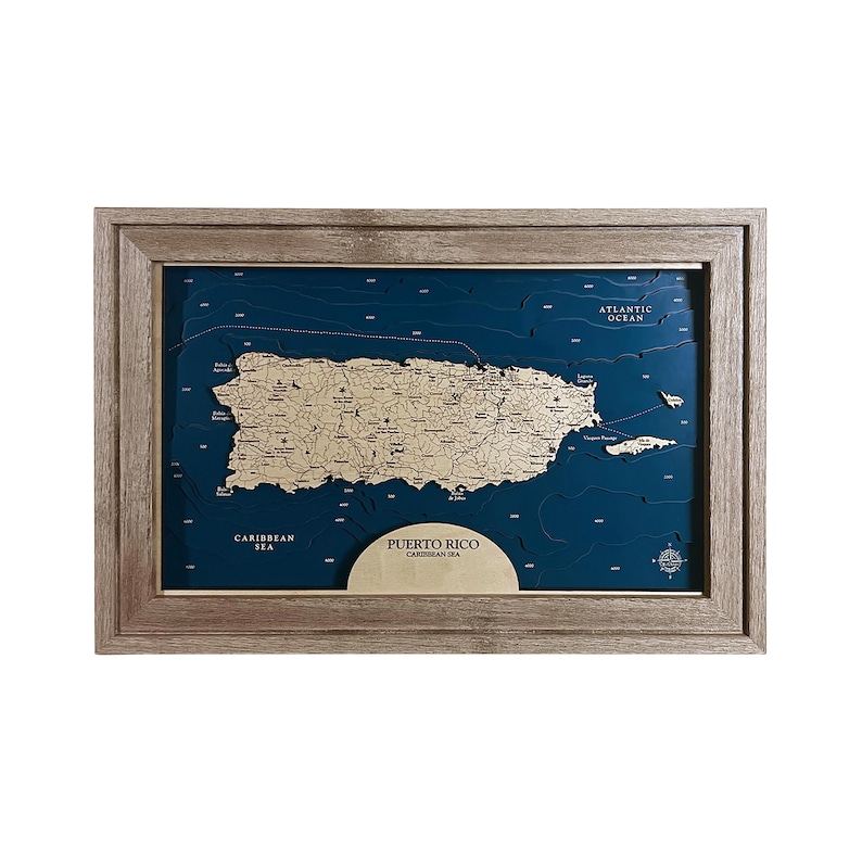 May include: A framed wooden map of Puerto Rico, featuring a gold-colored island against a deep blue background. The map includes the Caribbean Sea and Atlantic Ocean, with text indicating locations. The frame is a light brown color.