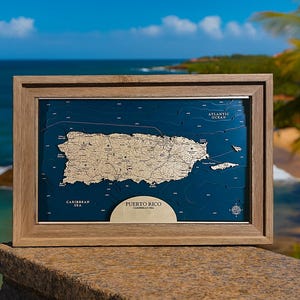 May include: A framed wooden map of Puerto Rico, featuring a detailed relief of the island in a light tan color against a deep blue background. The frame is a light brown color. The words "Puerto Rico" and "Caribbean Sea" are visible.