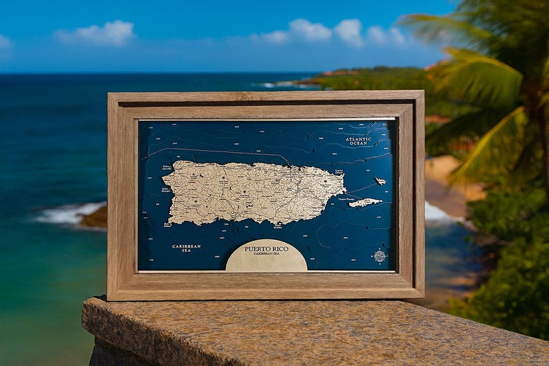 May include: A framed wooden map of Puerto Rico, featuring a detailed relief of the island in a light tan color against a deep blue background. The frame is a light brown color. The words "Puerto Rico" and "Caribbean Sea" are visible.