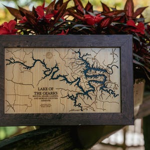 May include: A rectangular wooden framed map of the Lake of the Ozarks in Missouri. The light-colored wood surface features a dark blue laser-cut lake design, black road lines, a compass rose, and text detailing the counties and location.