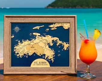 May include: A framed wooden map of St. John, U.S. Virgin Islands, featuring a tan landmass against a deep blue background with a compass rose and text labels. Beside it sits a tropical cocktail in a glass with a blue straw and pineapple slice.