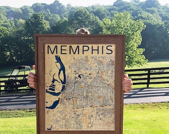 May include: A framed wooden map of Memphis featuring a light tan surface with intricate black street lines and a deep blue river section. The word MEMPHIS is printed in bold dark blue capital letters at the top of the rectangular wall art.