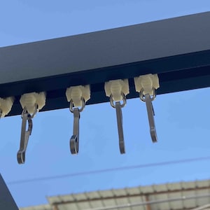 Op de afbeelding: Close-up van een zwarte metalen gordijnroede met verschillende crèmekleurige plastic glijders en metalen gordijnhaakjes. De haken zijn bevestigd aan de glijders, klaar om gordijnen te houden. De achtergrond is een heldere blauwe lucht.
