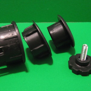 May include: Black plastic furniture leg components, including three cylindrical inserts and a threaded knob with a metal bolt. The parts are arranged on a green surface, suitable for furniture repair or construction.