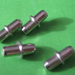 May include: Four silver-colored shelf support pins with a cylindrical body and a raised collar. The pins are designed to fit into pre-drilled holes in shelving units. The background is a solid green color.