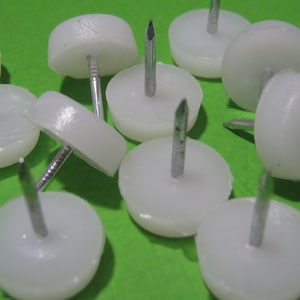 Può includere: Una collezione di feltrini per mobili in plastica bianca, ciascuno caratterizzato da una base liscia e arrotondata e da un chiodo metallico appuntito che sporge dal centro, progettato per essere fissato sotto le gambe dei mobili per facilitarne lo spostamento.
