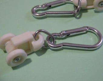 Può includere: Due cursori per binari per tende, ciascuno dotato di un carrello in plastica bianca con due piccole ruote e un moschettone in metallo argentato fissato a un anello metallico circolare. I componenti sono progettati per scorrere su un binario.