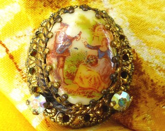 May include: A gold-toned brooch featuring a cameo with a painted scene of two figures in a garden. The cameo is surrounded by a decorative border of gold-toned metal and rhinestones.