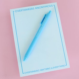 Pode incluir: Um bloco de notas azul claro com uma borda azul e o texto "Overthinkers Anonymous" no topo e "Overthinking Anything & Everything" na parte inferior. Uma caneta azul claro está sobre o bloco de notas.
