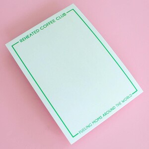 May include: A white notepad with a green border and the text "Reheated Coffee Club" and "Fueling Moms Around the World" printed on it.