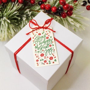 以下が含まれることがあります： 赤いリボンと「Good Tidings of Great Joy」と書かれた花柄のギフトタグが付いた白いギフトボックス。